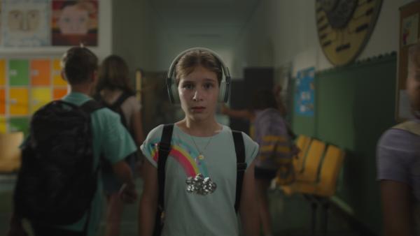 Eilen, gespielt von Julia Kovacs, steht mittig mit Kopfhörer in einem Schulgebäude. Um sie herum bewegen sich geschäftigt Personen mit Rucksäcken.  