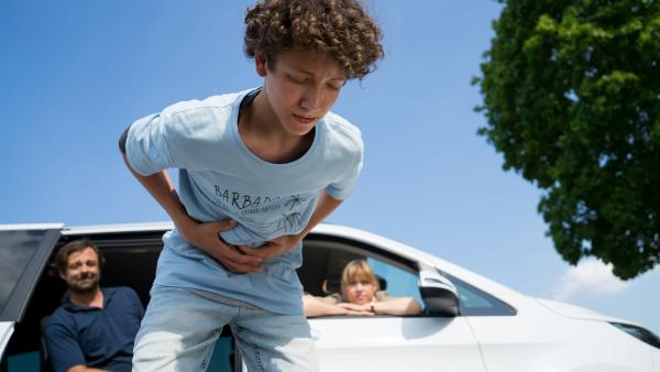Edvard steht nach vorne gebeugt vor einem Auto und hält mit schmerzvollem Gesichtsausdruck seine Hände am Bauch. Seine Eltern beobachten ihn aus dem Auto.