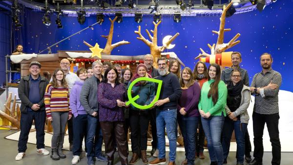 Eine große Menschengruppe im Fernsehstudio. Im Hintergrund befindet sich das KiKA-Baumhaus-Set. In der Mitte stehen Dr. Astrid Plenk und Matthias Franzmann und halten gemeinsam ein symbolisches Green Motion Label in den Händen.