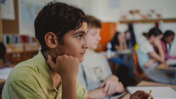 Luca (Baselius Göze) sitzt im Klassenzimmer am Tisch und schaut nachdenklich. Sein Kinn hat er auf seine Hand gestützt. Neben ihm sitzt Erik (Frederik Stein). Im Hintergrund sind noch weitere Kinder an ihren Schultischen.