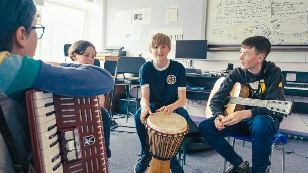 Calum und seine Freunde sind im Musikraum ihrer Schule. Sie sitzen mit ihren Instrumenten, Akkordeon, Trommel und Gitarre, im Halbkreis. Alle lächeln und scheinen sehr gut gelaunt zu sein.