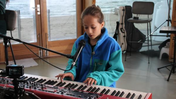 Ein Mädchen spielt auf einem E-Piano und schaut konzentriert auf die Tasten. Vor sich hat das Mädchen ein Mikrofon. Im Hintergrund ein Gitarrenkoffer und durch große Fensterscheiben ist ein Sandstrand und das Meer zu sehen.
