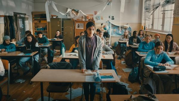 Mona steht in der Mitte in einem Klassenraum und schaut verunsichert auf eine aufgeschlagenes Buch, das vor ihr auf einem Tisch liegt. Links und rechts von ihr sitzen Jungen und Mädchen an ihren Schulbänken und schauen gelangweilt und genervt. Papier liegt auf dem Boden des Klassenzimmers, ein Poster hängt von der Wand heurnter und die Jalousien an den Fenstern hängen schief. 