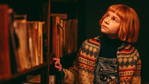 Nelly Rapp steht in einer dunklen Bibliothek und schaut sich die Bücherregale an.