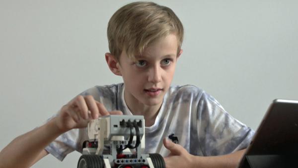 Friedrich sitzt am Tisch und hält ein kleines Fahrzeug in der Hand, das er programmiert hat. 