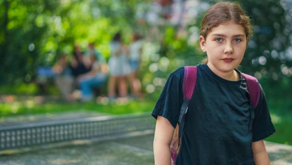 Lya (11) trägt ein schwarzes T-Shirt und einen lila Rucksack. Sie schaut ernst in die Kamera.