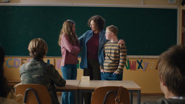 Eine Lehrerin steht mit Amanda und Lars vor der Tafel im Klassenzimmer. Sie schaut freudig zu Amanda, um ihr zu sagen, dass sie die Patenschaft für Lars übernimmt. 